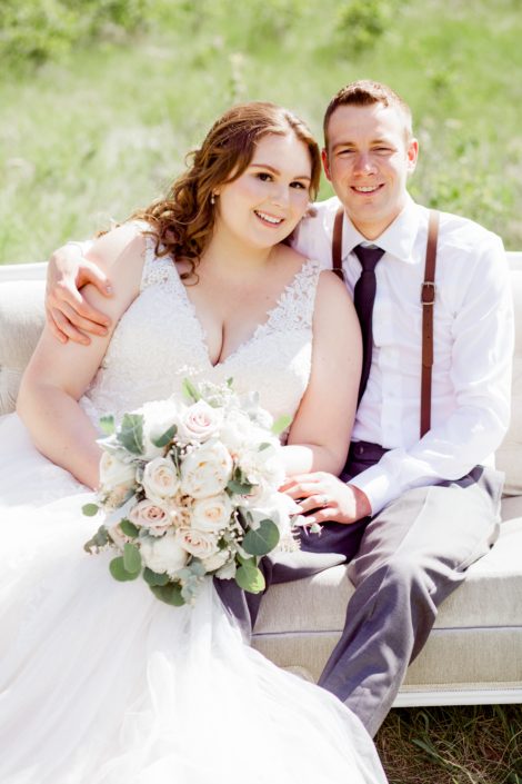 Kuera and Mark on a tufted sofa; Keura is holding a cream and blush bridal bouquet featuring white o'hara garden roses, quicksand roses, white peonies, pale pink astilbe, babies breath, dusty miller and eucalyptus; groom is wearing suspenders.