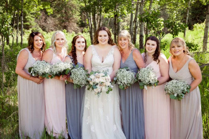 Kuera and her bridesmaids; bride is wearing a white lacey bridal gown and holding a cream and blush bouquet featuring white o'hara garden roses and white peonies with grey toned greenery; bridesmaids are wearing dusty blue, blush or grey floor-length dresses and holding babies breath bouquets with eucalyptus and dusty miller collars.