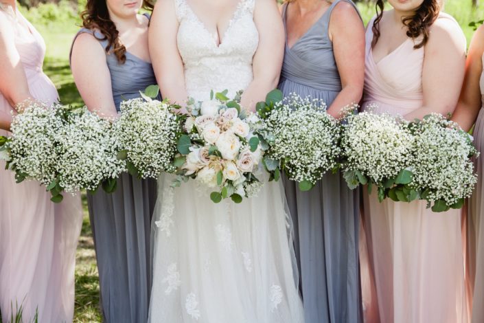 Close up of Kuera and her bridesmaid's vintage chic bouquets; Kuera is wearing a lacey white gown and holding a cream and blush bouquet featuring white peonies, white o'hara garden roses, quicksand roses, pale pink astilbe, babies breath, dusty miller and eucalyptus; bridesmaids are wearing dusty blue, blush or grey dresses and holding babies breath bouquets with eucalyptus and dusty miller collars.