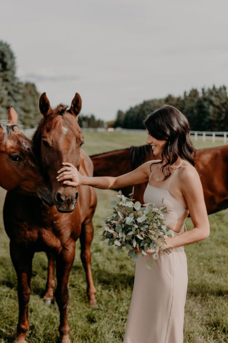 Erika' bridesmaid wearing mauve dress and holding a fresh mixed eucalyptus bouquet and petting a horse.