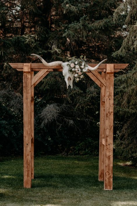 Wooden archway with longhorn skull attached at the centre decorated with quicksand roses, white ranunculus, ivory lisianthus and panda anemones with fresh mixed eucalyptus greenery.