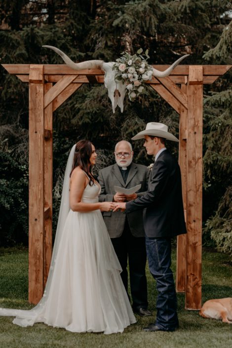 Erika and Colt at their wedding ceremony standing under the wooden archway with longhorn skull at the centre that is decorated with quicksand roses, white lisianthus, panda anemones, ranunculus and eucalyptus greenery.