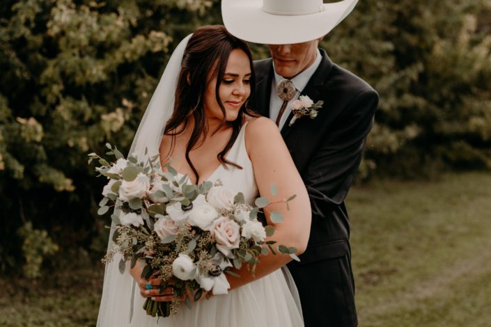 Colt and Erika embracing - colt is wearing a blush and white boutonniere made with spray roses and eucalyptus. Erika is holding a blush and white bridal bouquet featuring quicksand roses, ivory ranunculus, panda anemones, lisianthus and a variety of eucalyptus greenery.