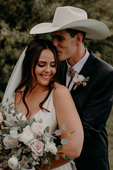 Colt kissing Erika. The groom is wearing a blush and white boutonniere made of spray roses and eucalyptus. The bride is holding a blush, ivory and white bouquet featuring quicksand roses, ranunculus, panda anemones, and lisianthus with a mixed variety of eucalyptus greenery.
