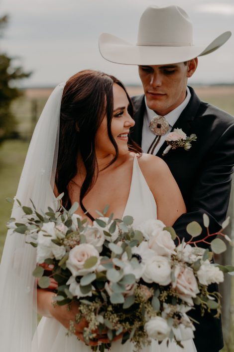 Erika and Colt embracing; groom is wearing a blush and white boutonniere made of roses and eucalyptus; bride is holding a blush, ivory and white bridal bouquet featuring quicksand roses, panda anemones, lisianthus, ranunculus and a mixed variety of eucalyptus greenery.