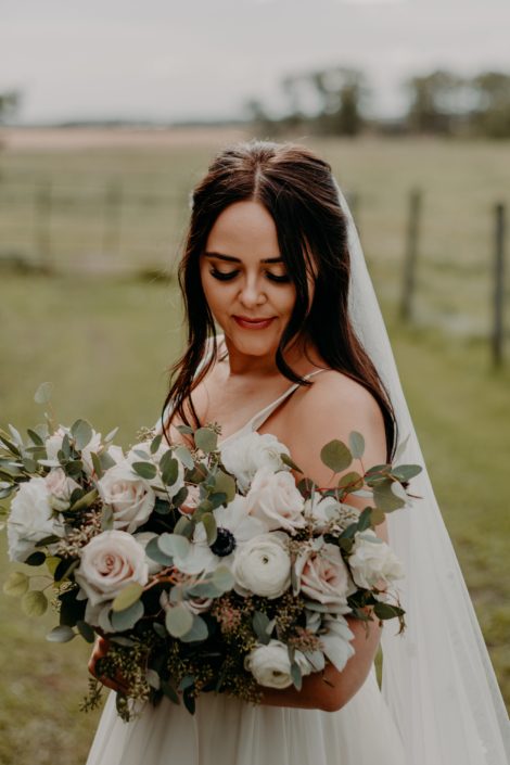 Bride Erika looking at her blush, ivory and white bridal bouquet featuring quicksand roses, ranunculus, panda anemones, lisianthus and mixed eucalyptus.