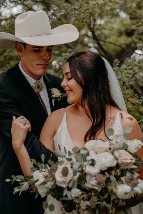Erika and Colt's Blush and Mauve Country Wedding - bride and groom embracing; groom is wearing a cowboy hat and blush and ivory boutonniere made of spray roses; bride is holding a blush, ivory and white bridal bouquet featuring quicksand roses, lisianthus, panda anemones and ranunculus with fresh eucalyptus.