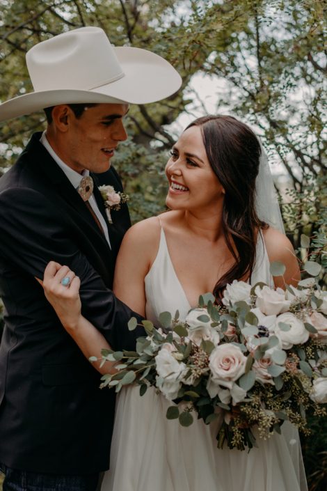 Bride and groom smiling at one another; bride is holding a blush and ivory bridal bouquet featuring panda anemones, lisianthus, quicksand roses and ranunculus with fresh eucalyptus greenery.