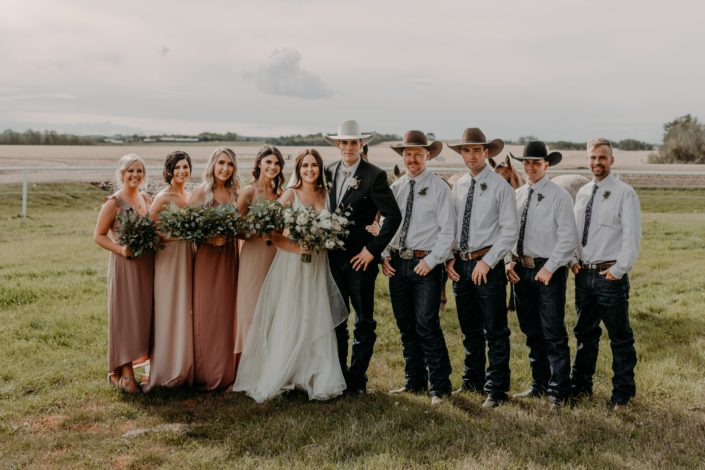 Erika and Colt's Blush and Mauve Country Wedding bridal party. The bridesmaids are wearing mauve floor length dresses carrying eucalyptus bouquets, the groom and groomsmen are wearing cowboy hats and boutonnieres and the bride is wearing a white dress and carrying a white, ivory and blush bouquet.