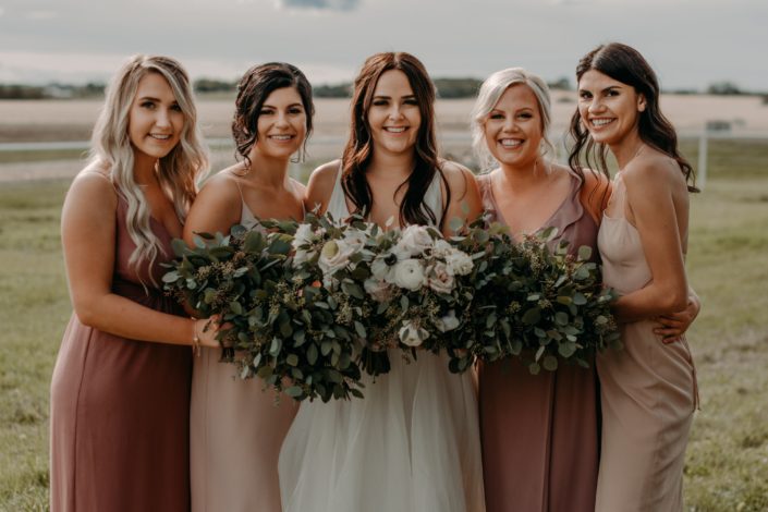 Erika and her bridesmaids - the bridesmaids are wearing mauve dresses and carrying fresh mixed eucalyptus bouquets and the bride is wearing a white bridal gown and carrying a white, ivory and blush bouquet featuring quicksand roses, panda anemones, white ranunculus and white lisianthus with eucalyptus greenery.