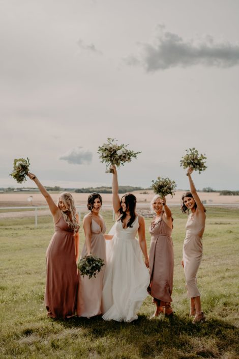 Bride and bridesmaids having fun and holding their bouquet in the air. The bridesmaids are wearing mauve dresses and carrying fresh mixed eucalyptus bouquets and Erika is wearing a white bridal gown and holding a blush, ivory and white bouquet with eucalyptus greenery.