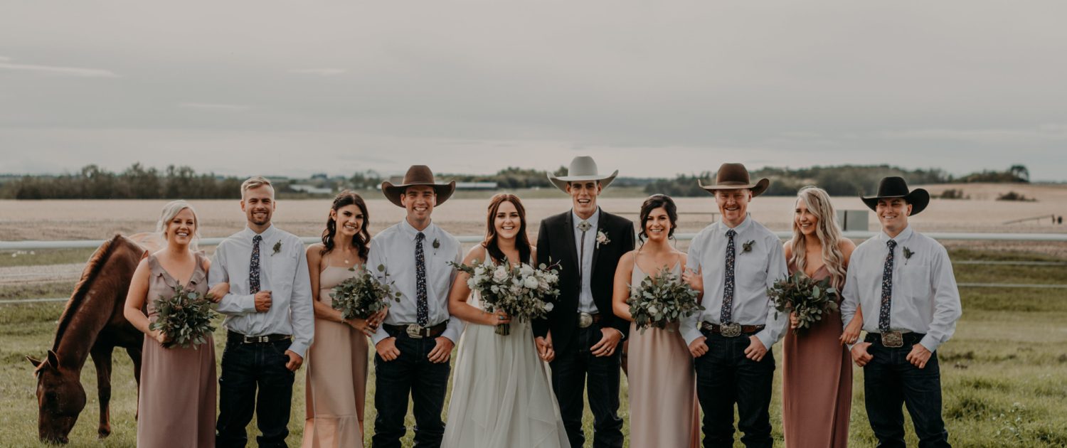 Erika and Colt's blush and mauve country wedding bridal party. The groom and his groomsmen are wearing cowboy hats and boutonnieres, the bridesmaids are wearing mauve dresses and carrying fresh mixed eucalyptus bouquets and the bride is wearing white and holding a blush, ivory and white bridal bouquet featuring roses, ranunculus, anemones and lisianthus.