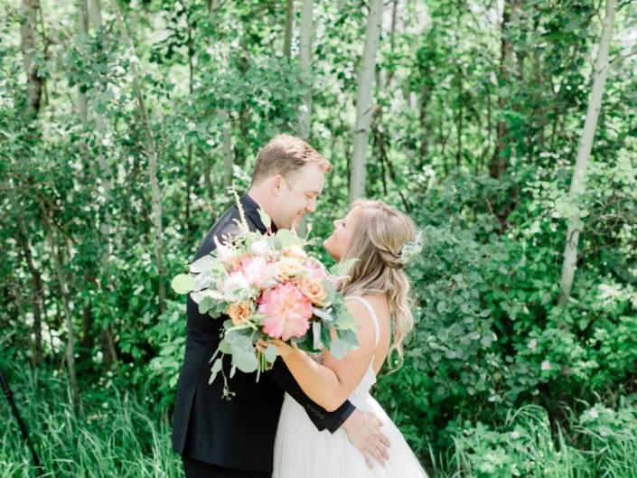 Taylor and Griffin smiling at each other holding lush and modern bouquet featuring coral charm peonies, quicksand roses, golden mustard roses, astilbe, astrantia, cappuccino roses, monstera leaves, eucalyptus and boston fern.