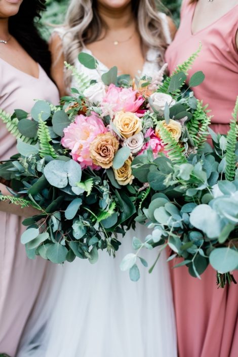 Bridal bouquet made of coral charm peonies, golden mustard roses, quicksand roses, cappuccino roses, astilbe, astrantia, boston fern, monstera leaf and a mixed variety of eucalyptus amongst bridesmaids bouquets made of eucalyptus and boston fern.