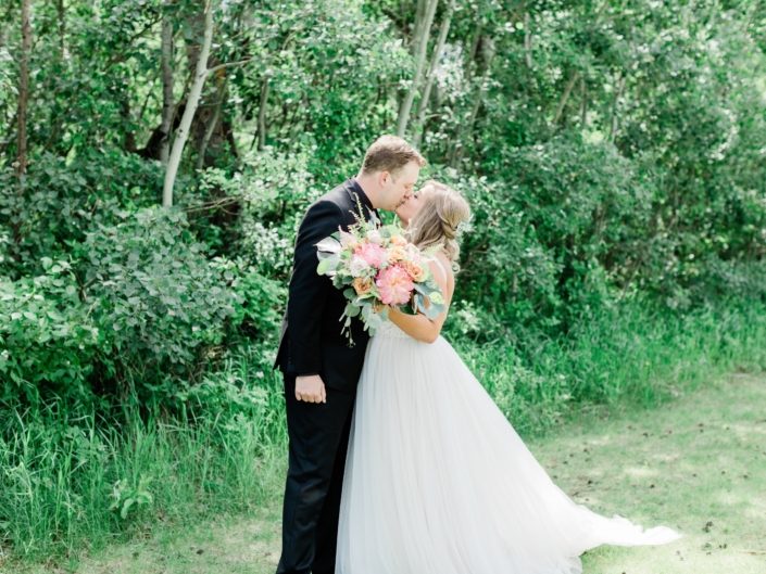 Talyor and Griffin kissing while holding a bouquet made of coral charm peony, yellow roses, monstera leaf, boston fern, and eucalyptus greenery.