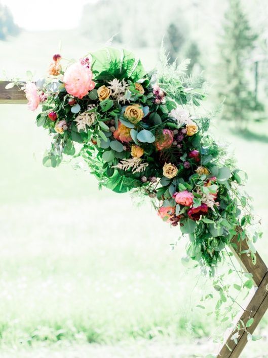 Modern Coral Charm Peony arrangement on a wooden hexagon arch featuring coral charm peonies, golden mustard roses, cappuccino roses, burgundy dahlias, astrantia, astilbe, plumosa, monstera leaves, and eucalyptus greenery.
