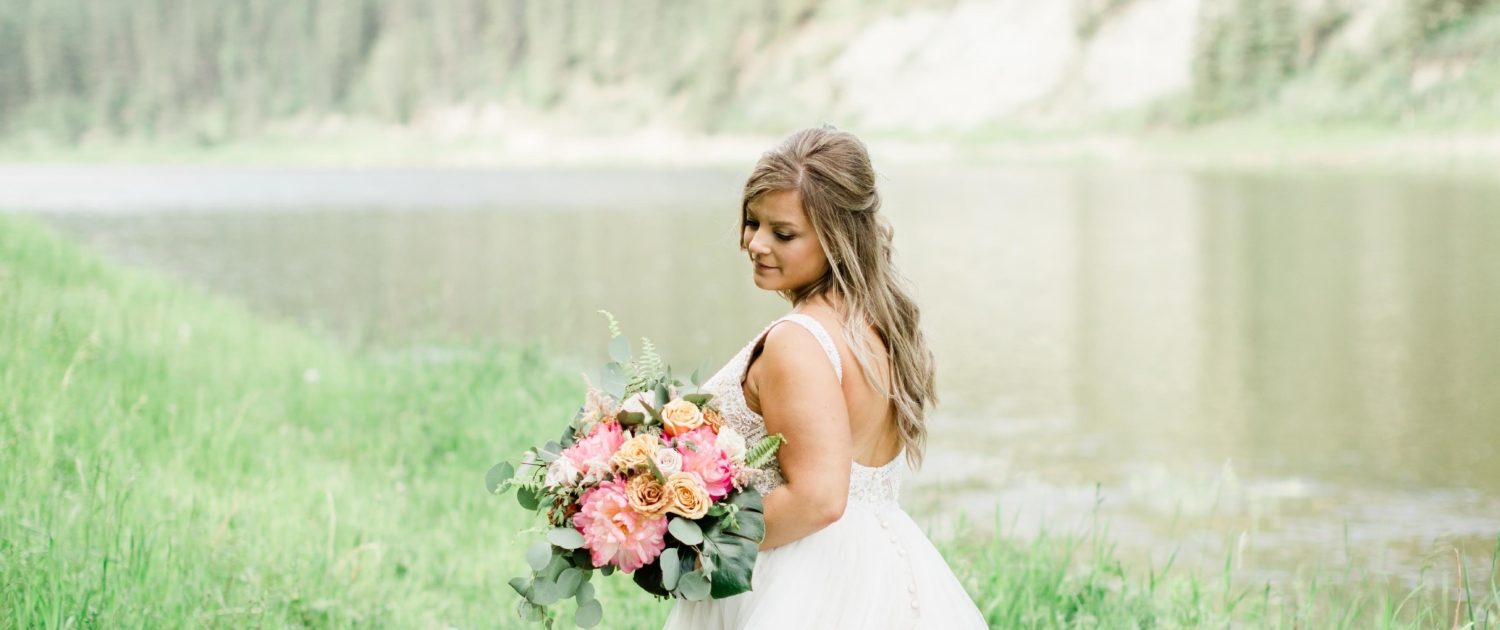 Bride Taylor wearing a white bridal gown and holding a coral charm peony bouquet with golden mustard , quicksand and cappuccino roses, monstera leaves, boston fern and eucalyptus greenery.