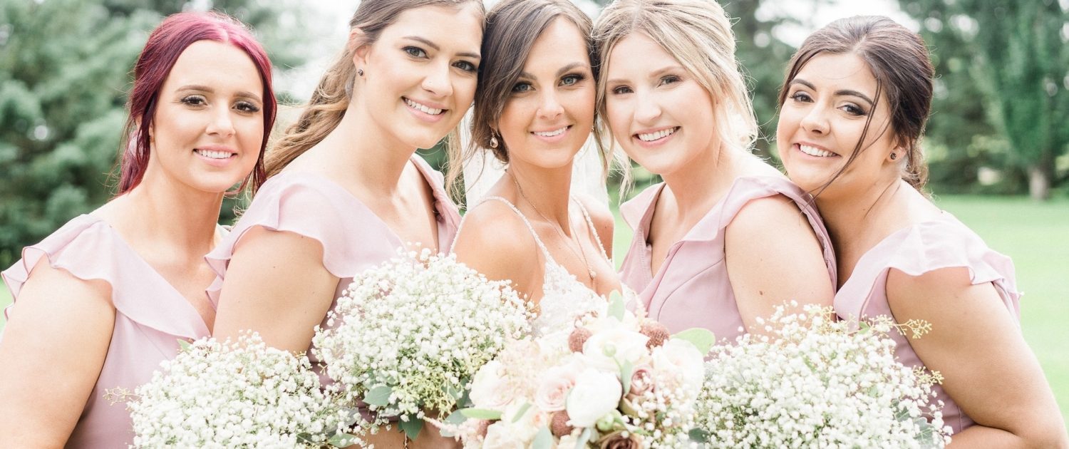 Bride, Taryn, surrounded by her bridesmaids; bride is wearing a white lace bridal gown and holding a dusty rose bouquet featuring rose gold painted scabiosa pods, white o'hara garden roses, quicksand and amnesia roses, ranunculus, astilbe, babies breath, dusty miller and eucalyptus greenery; bridesmaids are wearing elegant rustic dusty pink dresses and carrying babies breath bouquets.