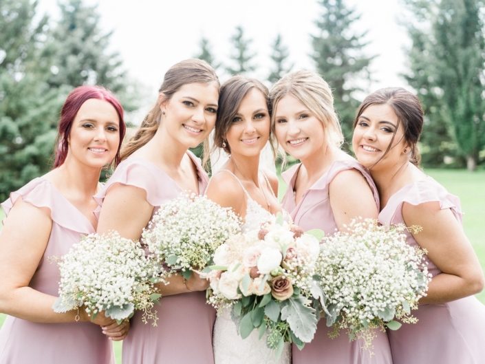 Bride, Taryn, surrounded by her bridesmaids; bride is wearing a white lace bridal gown and holding a dusty rose bouquet featuring rose gold painted scabiosa pods, white o'hara garden roses, quicksand and amnesia roses, ranunculus, astilbe, babies breath, dusty miller and eucalyptus greenery; bridesmaids are wearing elegant rustic dusty pink dresses and carrying babies breath bouquets.