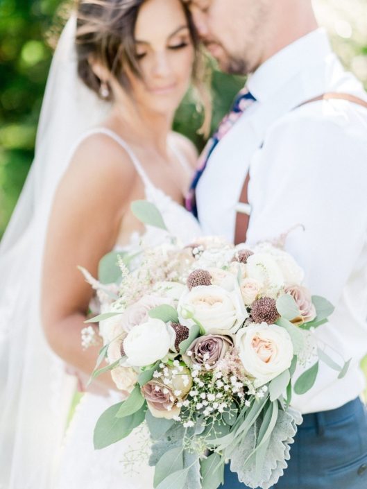 Bride standing with groom holding bouquet made with rose gold painted scabiosa pods, dusty rose quicksand and amnesia roses, blush white o'hara garden roses, white ranunculus, pink astilbe, babies breath, and grey toned greenery such as dusty miller and eucalyptus.