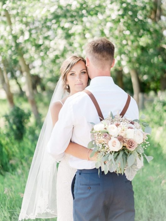 Bride hugging groom and holding bouquet designed with rose gold painted scabiosa pods, white o'hara garden roses, dusty pink amnesia and quicksand roses, astilbe, raununculus, babies breath, dusty miller and eucalyptus.