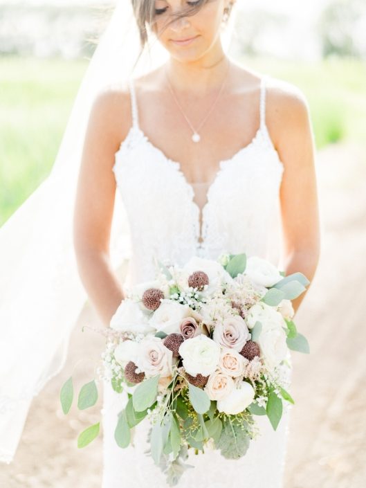 Bride wearing white lace dress and holding a dusty rose bridal bouquet featuring rose gold painted scabiosa pods, white o'hara garden roses, quicksand roses, amnesia roses, astilbe, babies breath, ranunculus, dusty miller and eucalyptus grey toned greenery.