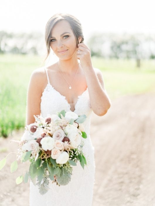 Bride, Taryn, wearing a white lace gown and holding a bouquet made with dusty pink amnesia and quicksand roses, white o'hara garden roses, rose gold painted scabiosa pods, ranunculus, astilbe, babies breath, dusty miller, and eucalyptus.