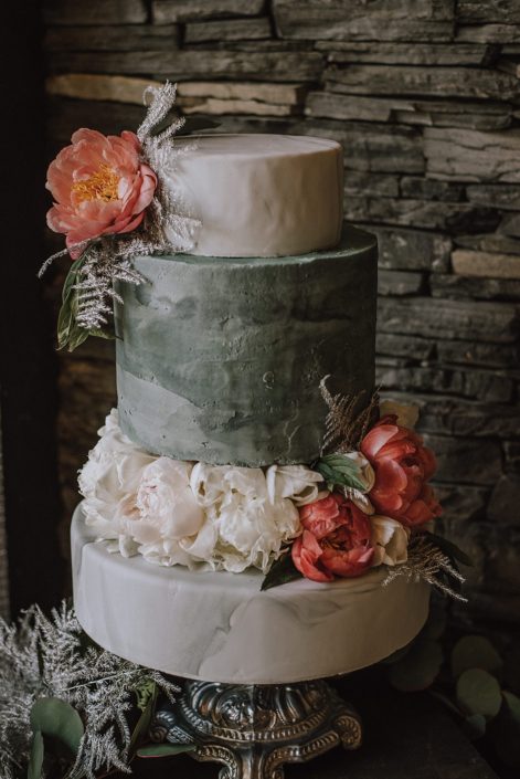 Tiered cake by Stella Bean Sweets for the Canyon Ski Resort Open House 2019. Cake on a marble stand and is adorned with coral charm and white peonies with silver painted plumosa.