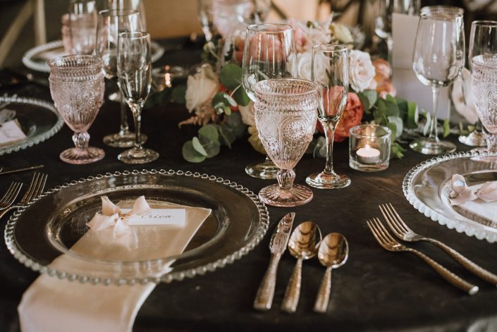 Place setting with clear and blush tableware, silverware, stationary, linens and a low coral and blush flower arrangement featuring roses and coral charm peonies.