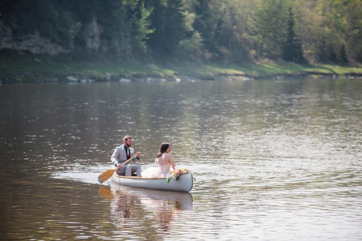 Bride and groom in a canoe on the river. A coral and blush bouquet is laying across the front of the canoe.
