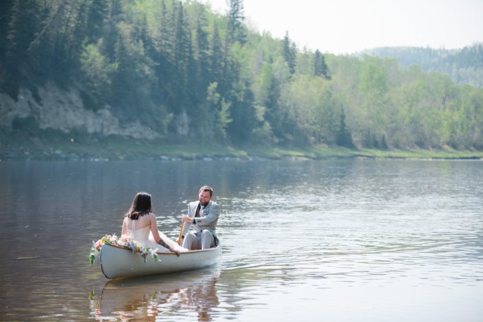 Bride and groom in a canoe on the river. The canoe is adorned with a blush and coral bouquet.