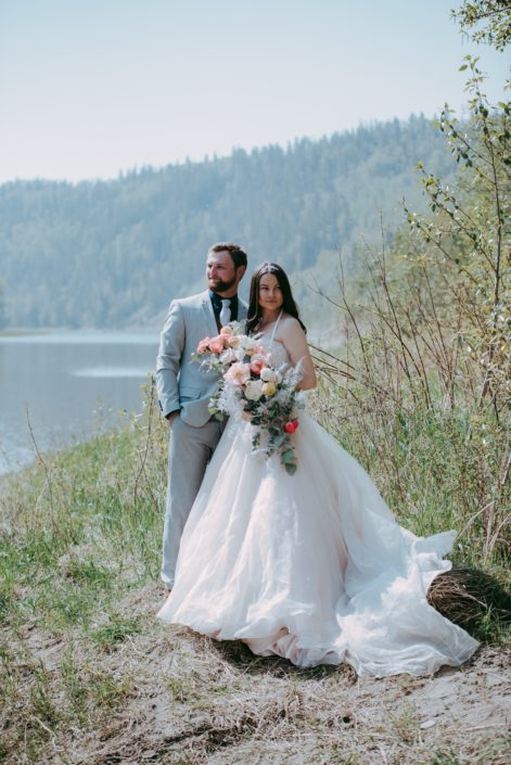Bride and groom standing beside a river holding a crescent shaped bouquet featuring coral charm peonies, quicksand and playa blanca roses, peach ranunculus, pale pink astilbe, silver plumosa and a mixed variety of eucalyptus greenery.