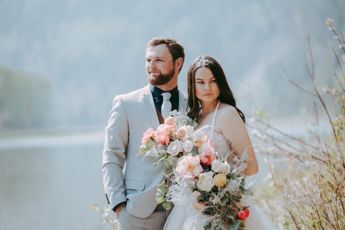 Bride and groom with a crescent shaped bouquet designed with coral charm peonies, quicksand and playa blanca roses, peach ranunculus, pale pink astilbe, metallic silver plumosa and eucalyptus greenery.