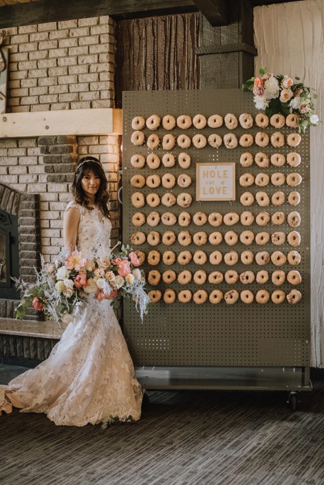 Bride holding a large blush and coral bouquet featuring coral charm peonies, roses, silver plumosa and greenery standing next to a doughnut wall with a floral arrangement attached to the top corner.