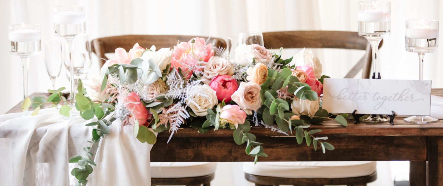 Sweetheart table arrangement designed with coral charm peonies, quicksand roses, playa blanca roses, peach ranunculus, light pink astilbe, silver plumosa, salal and a variety of eucalyptus greenery.