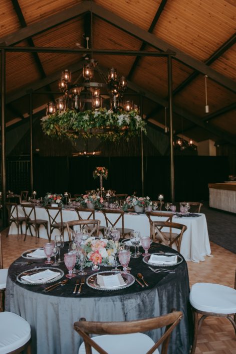 Reception hall at Canyon Ski Resort for the Open House 2019; greenery garland on chandelier; round table with grey table cloth, blush goblets and vineyard chairs; rectangle tables with ivory tablecloths, blush goblets, silver lucca flatware, rustic vineyard chairs; floral arrangement designed with coral charm peonies, blush roses and eucalyptus greenery.