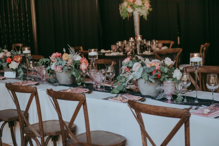 Rectangular table decorated with an ivory velvet table cloth, smoked grey table runner, blush goblets, and coral and blush floral arrangement in concrete vases featuring coral charm peonies, quicksand roses, playa blanca roses, peach ranunculus, silver plumosa and eucalyptus; vineyard chairs.