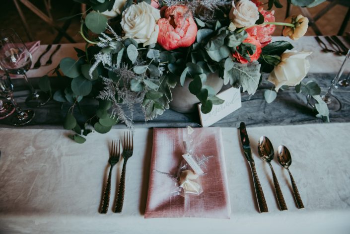Tablescape for Canyon Ski Resort Open House 2019; ivory velvet tablecloth, blush napkin, fortune cookie, silver lucca flatware, smoked grey velvet table runner, and a coral and blush floral arrangement in a concrete vase designed with coral charm peonies, quicksand roses, playa blanca roses, peach ranunculus, silver plumosa, and a mixed variety of eucalyptus greenery.