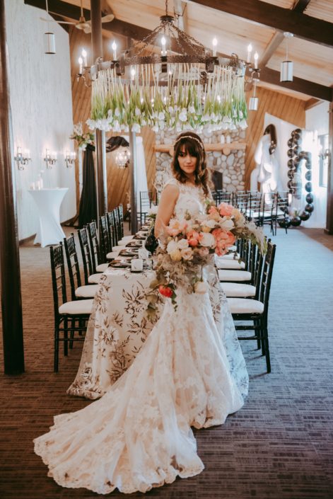 Bride wearing lace gown and holding a crescent shaped bouquet designed with coral charm peonies, blush roses, peach ranunculus, silver plumosa and eucalyptus greenery; standing in front of a black and white decorated rectangle table with a tulips hanging from the chandelier at Canyon Ski Resort Open House 2019.