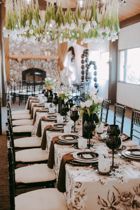 Rectangular table decorated with an ivory and black marie noir linen tablecloth, black and white plates, black goblets and black chiavari chairs; black vases villed with toffee and quicksand roses, white tulips and mazanita branches; tulips hanging from the chandelier.