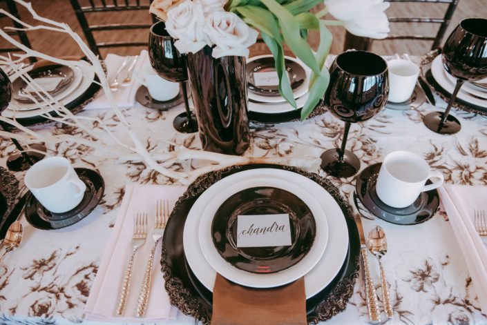 Place setting for Canyon Ski Resort Open House 2019 featuring black and ivory marie noir linen tablecloth, black and white plates, black goblets, and black vases filled with quicksand and toffee roses and white dream tulips; manzanita branches laying on table.