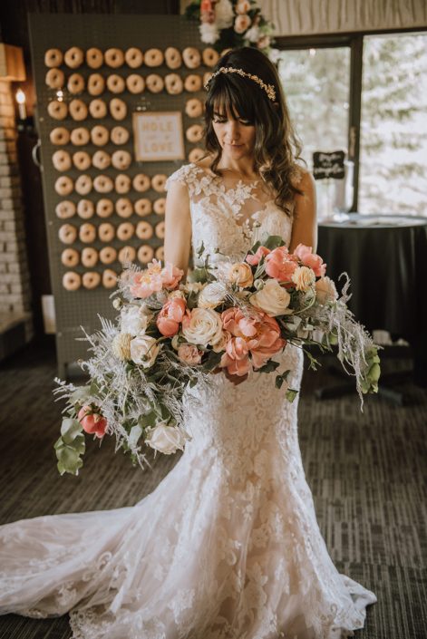 Bride wearing a lace gown from Novia Mia Bridal and holding a crescent shaped bouquet featuring coral charm peonies, peach ranunculus, quicksand roses, playa blanca roses, pale pink astilbe, silver painted plumosa and a mixed variety of eucalyptus greenery.