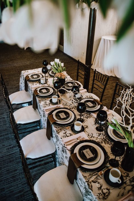Rectangular table set with an ivory and black marie noir linen tablecloth, black and white plates, black goblets, and black chiavari chairs; black vases were filled with white dream tulips, toffee and quicksand roses and Mazanita branches.