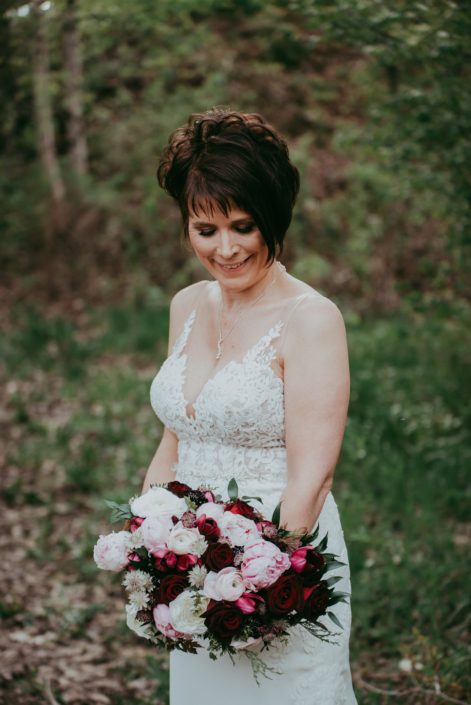 Bride looking down at her elegant pink and burgundy bouquet made with Sarah Bernhardt peonies, burgundy Helleborus, blush and white ranunculus, black bacarra roses, blackberry scoop scabiosa, burgundy tulips, pale pink astrantia and eucalyptus.