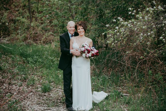 Bride and groom, Sandra and David, embracing and holding an elegant pink and burgundy bouquet designed with Sarah Bernhardt peonies, Helleborus, Ranunculus, Black Bacarra roses, Blackberry Scoop Scabiosa, Burgundy Tulips, Pale Pink Astrantia and eucalyptus.