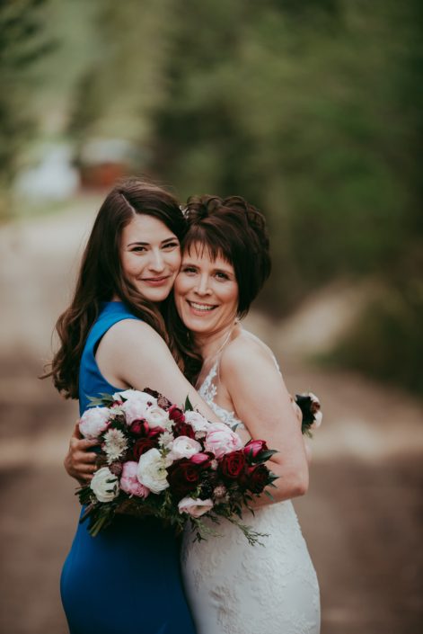 Bride hugging daughter holding a pink and burgundy bridal bouquet featuring burgundy helleborus, Sarah Bernhardt peonies, Blush and White ranunculus, black baccara roses, blackberry scoop scabiosa, burgundy tulips and accented with pale pink astrantia and seeded eucalyptus greenery.