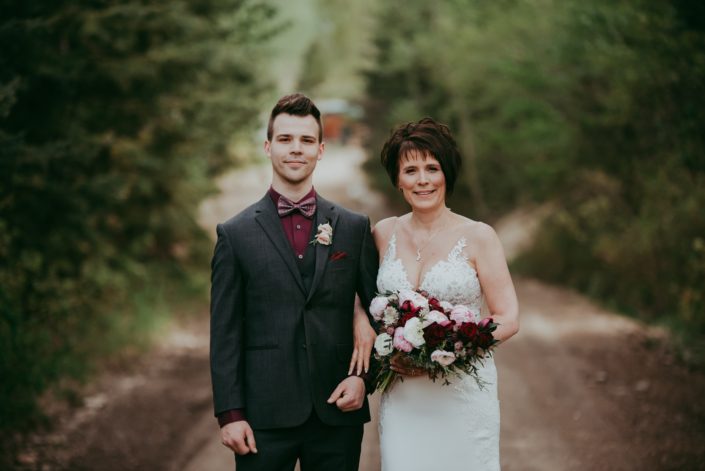 Bride and Son; Bride holding a pink and burgundy bridal bouquet featuring Sarah Bernhardt peonies, blush and white ranunculus, black baccara roses, blackberry scoop scabiosa, burgundy tulips and accented with pale pink astrantia and seeded eucalyptus greenery; son wearing a boutonniere made with blush spray roses and burgundy ninebark leaves and juniper.