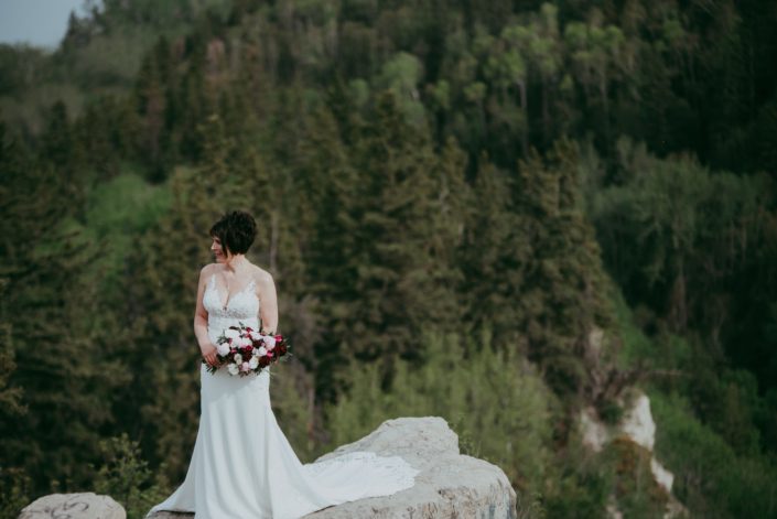 Bride, Sandra, standing on a cliff and holding her pink and burgundy bouquet designed with helleborus, peonies, ranunculus, black bacarra roses, blackberry scoop scabiosa, tulips, astrantia and eucalyptus.