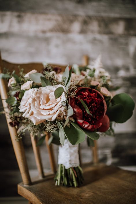 Rustic red and blush bridal bouquet designed with quicksand roses, red charm peony, burgundy astrantia, pale pink astilbe, and blush spray roses finished with eucalyptus greenery and a blush satin with lace overlay handle wrap.