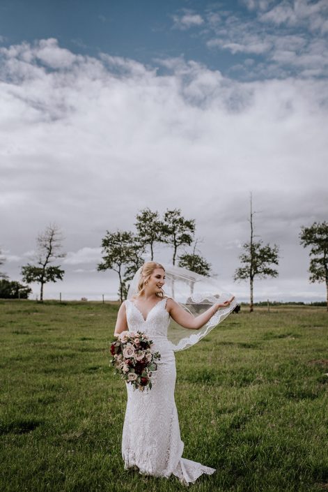 Bride standing in a field fanning her veil and holding a rustic red and blush bridal bouquet featuring quicksand roses, red charm peonies, blush spray roses, burgundy astrantia, light pink astilbe and eucalyptus greenery.